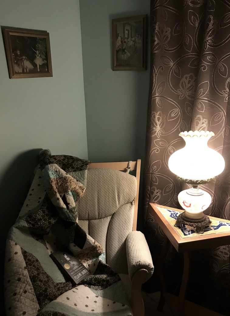 Chair in corner with quilt, book and a side table with antique reading lamp