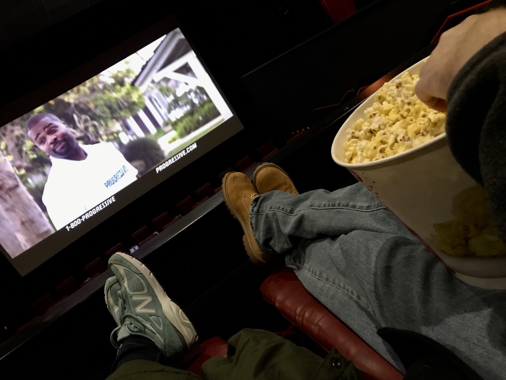 bucket of popcorn on 2 pairs of legs with movie screen in background