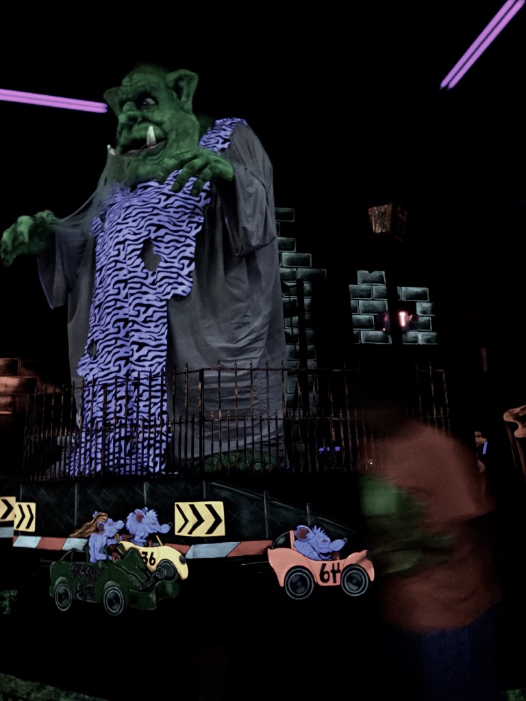Big green orc looking monster lit with black lights and a blurry kid in the foreground that looks like a ghost.
