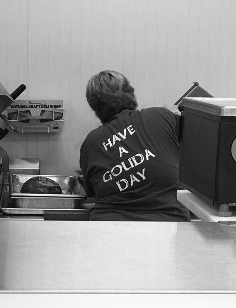 Women slicing lunch meat and the back of her shirt says "Have a Gouda Day".